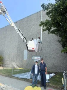 Two veterans stand in front of a flagpole with painters in the raised bucket of a firetruck's ladder behind them.